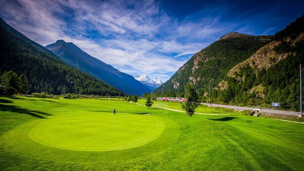 A golf course with a red train in the background, surrounded by mountains and clear skies. - Haus Bergdohle (Randa)