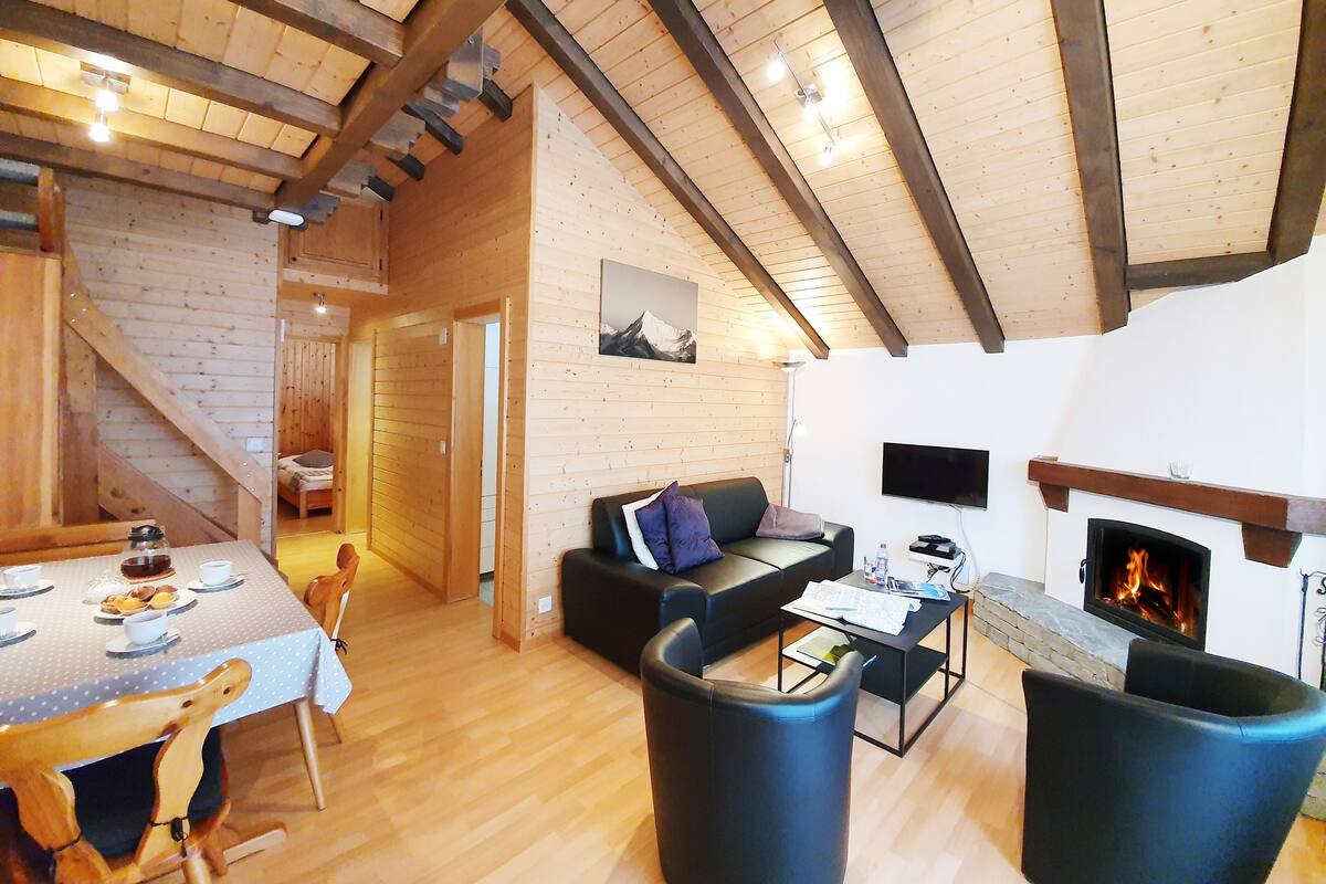 A cozy cabin interior with a dining area, a sofa, a fireplace, and a staircase leading to another room.