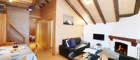 A cozy cabin interior with a dining area, a sofa, a fireplace, and a staircase leading to another room.