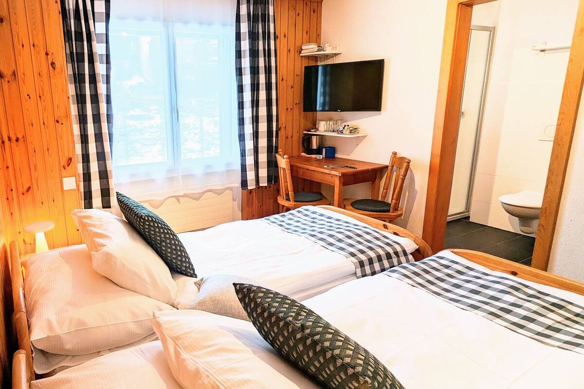 A hotel room with two beds, a desk, a chair, a TV, and a bathroom visible through an open door.