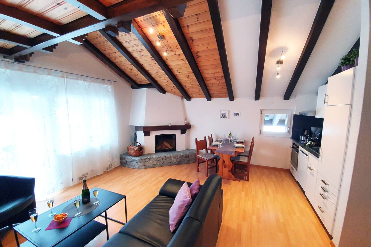 A cozy living room with a fireplace, wooden beams, a dining area, and a kitchen.