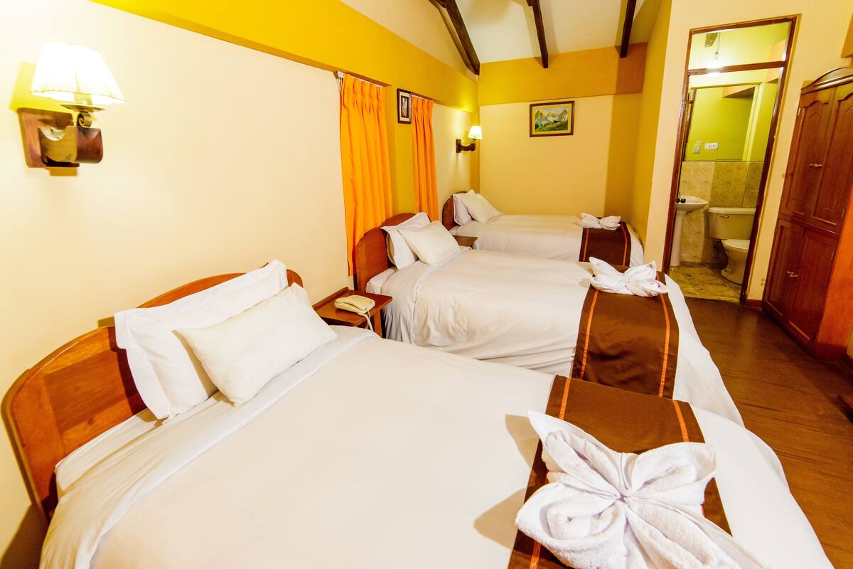 Une chambre d’hôtel avec trois lits, chacun recouvert de draps blancs et muni d’une serviette. On y trouve une tête de lit en bois sur le côté gauche, une table de chevet avec une lampe et un tableau accroché au mur.