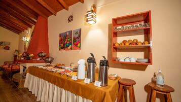 Disposição de buffet com uma mesa coberta por um tecido amarelo, diversos tipos de comida e uma estação de café com duas chaleiras elétricas.