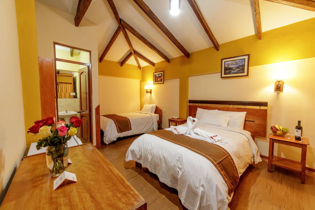 Une chambre d’hôtel avec deux lits, des poutres en bois au plafond, une table en bois ornée d’un vase de fleurs et une petite table sur laquelle se trouvent une bouteille et des fruits.