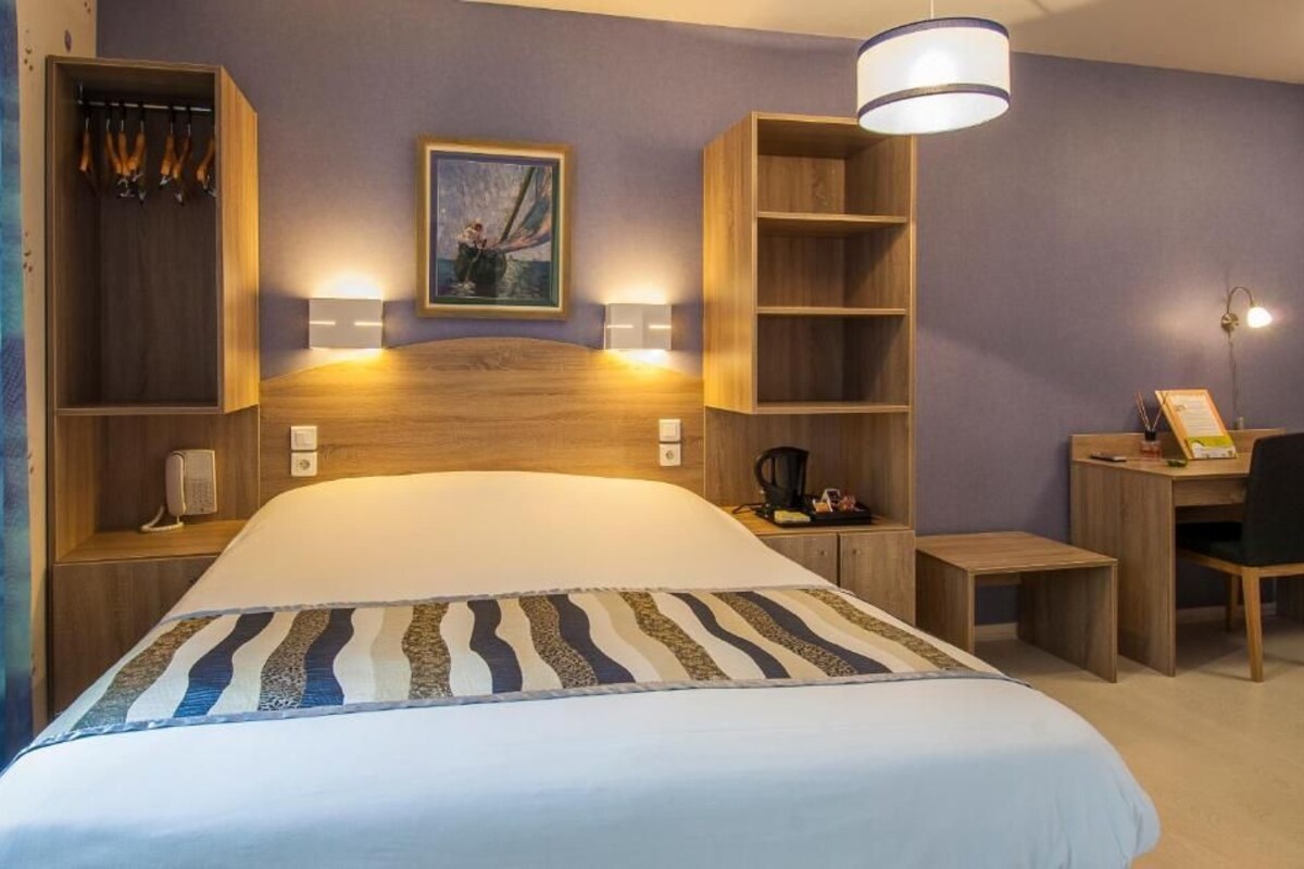 A hotel room with a large bed, a desk, a chair, a coat rack, and a painting on the wall.
