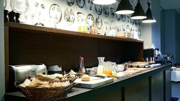 A breakfast buffet area with various pastries and bread, a wall with black and white portraits, and pendant lighting. - Square Hotel (Kortrijk)