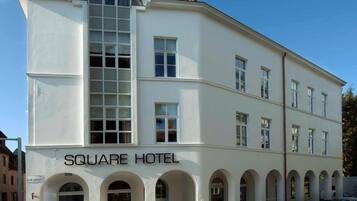 Un bâtiment blanc avec une enseigne « SQUARE HOTEL » sur la façade. Il possède de nombreuses fenêtres et une partie incurvée à l’étage supérieur.