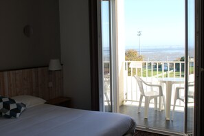 A hotel room with a bed, a nightstand, a lamp, and a view of a balcony with chairs and a table. - Hotel Mer et Forêt (Saint-Trojan-les-Bains)
