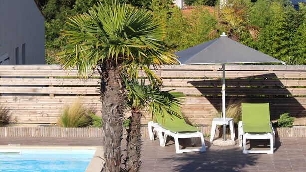 A swimming pool with lounge chairs and a palm tree, surrounded by a wooden fence and greenery. - Hotel Mer et Forêt (Saint-Trojan-les-Bains)