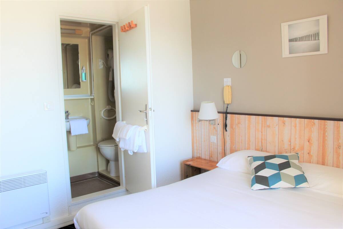 Ein Hotelzimmer mit Bett, Nachttisch mit Lampe, Holz-Kopfstütze, Bad mit Toilette und Handtuchhalter sowie einem an der Wand befestigten Telefon.