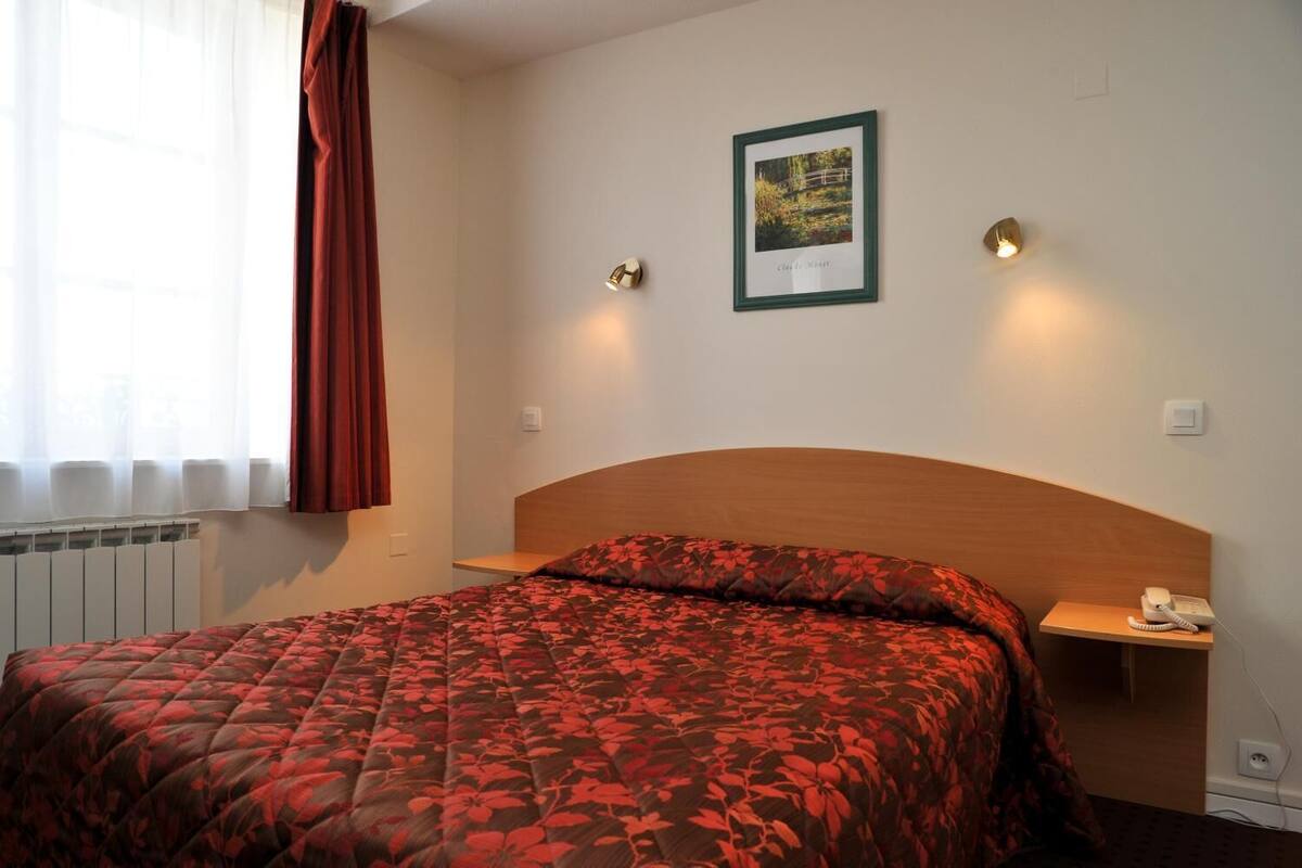 Une chambre d’hôtel avec un lit, une table de chevet avec un téléphone, un tableau encadré au mur et une fenêtre avec des rideaux.