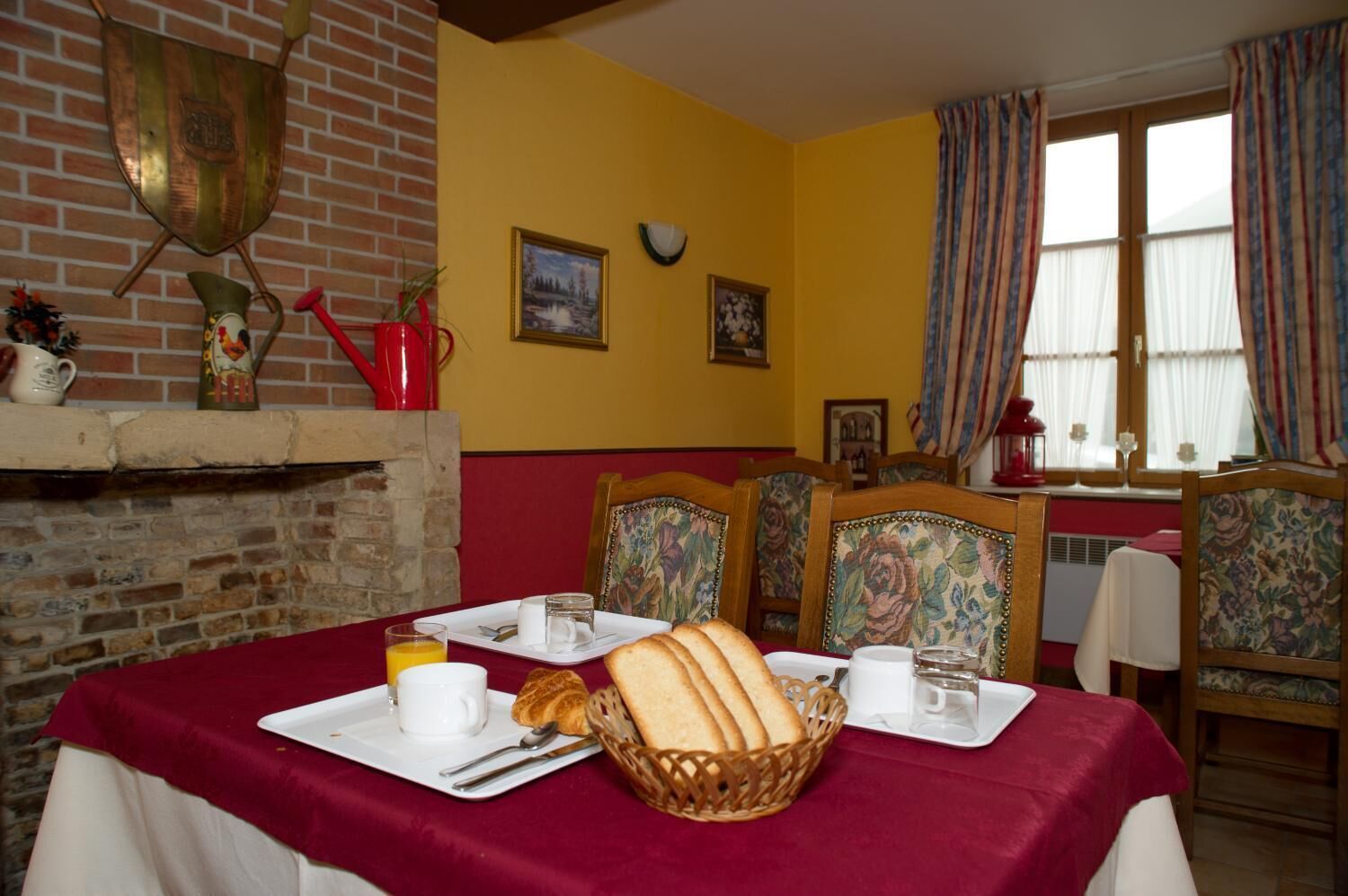 A dining area with a brick fireplace, yellow walls, floral chairs, and a table set with bread, plates, and glasses.