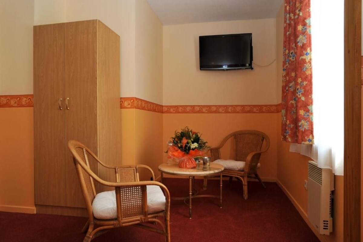 Una habitación con armario, dos sillas de mimbre, una mesita con un centro de mesa floral, un televisor montado en la pared y un radiador.
