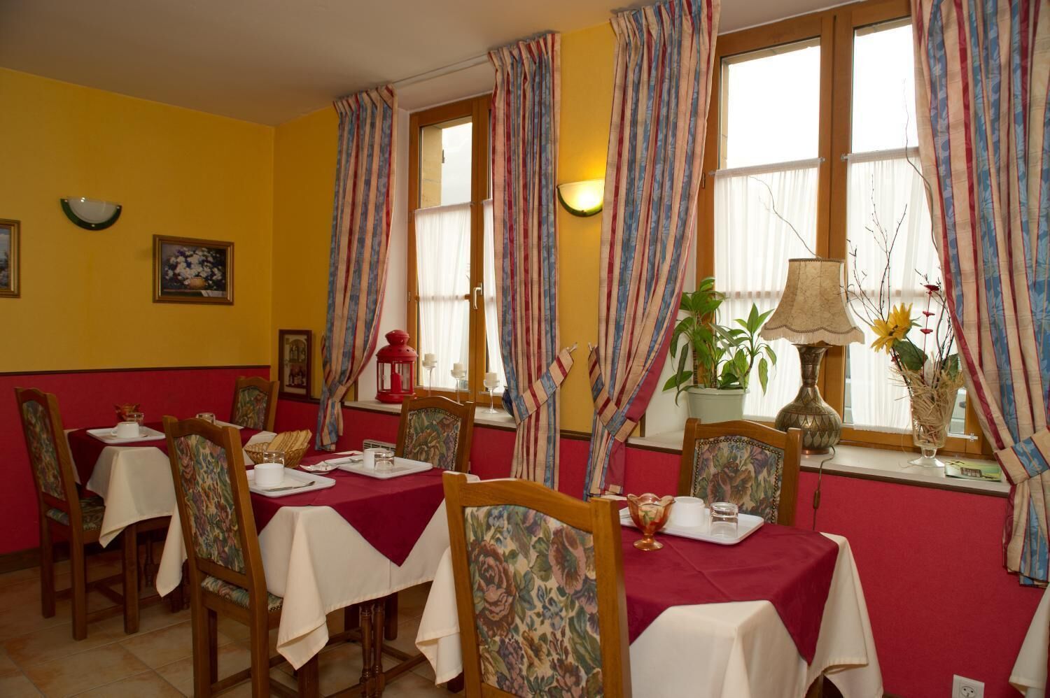 A dining area with tables set for four, each with a red tablecloth and white napkins. There are chairs with floral upholstery. The room has yellow walls, large windows with curtains, and a potted plant on the windowsill.