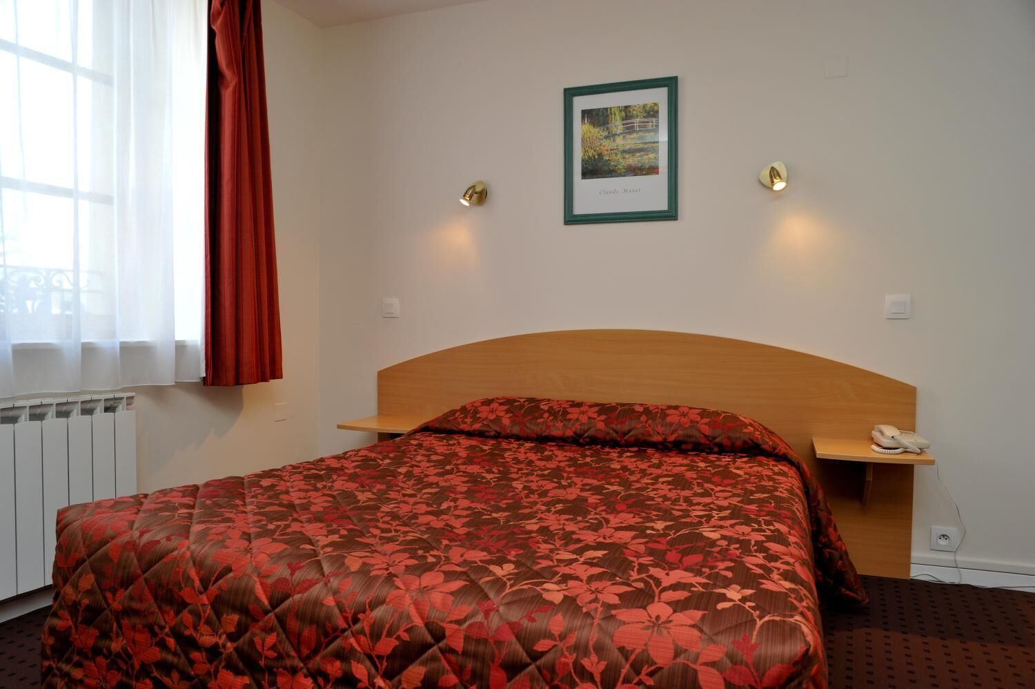 A hotel room with a bed, a bedside table with a phone, a framed picture on the wall, and a window with curtains.