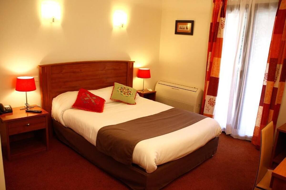 Habitación de hotel con cama de madera, dos mesitas de noche con lámparas, un radiador y una ventana con cortinas.