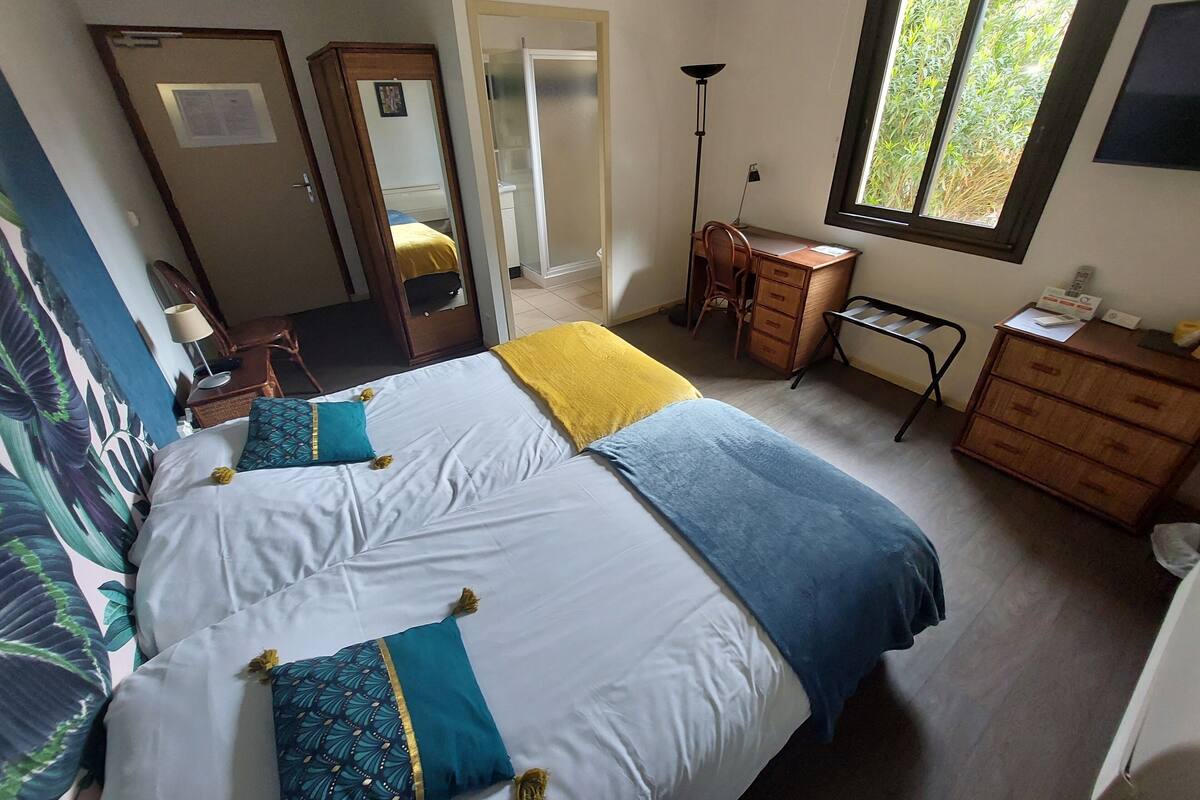 Un dormitorio con dos camas, un escritorio, una silla, un armario y una ventana con vistas a la vegetación.