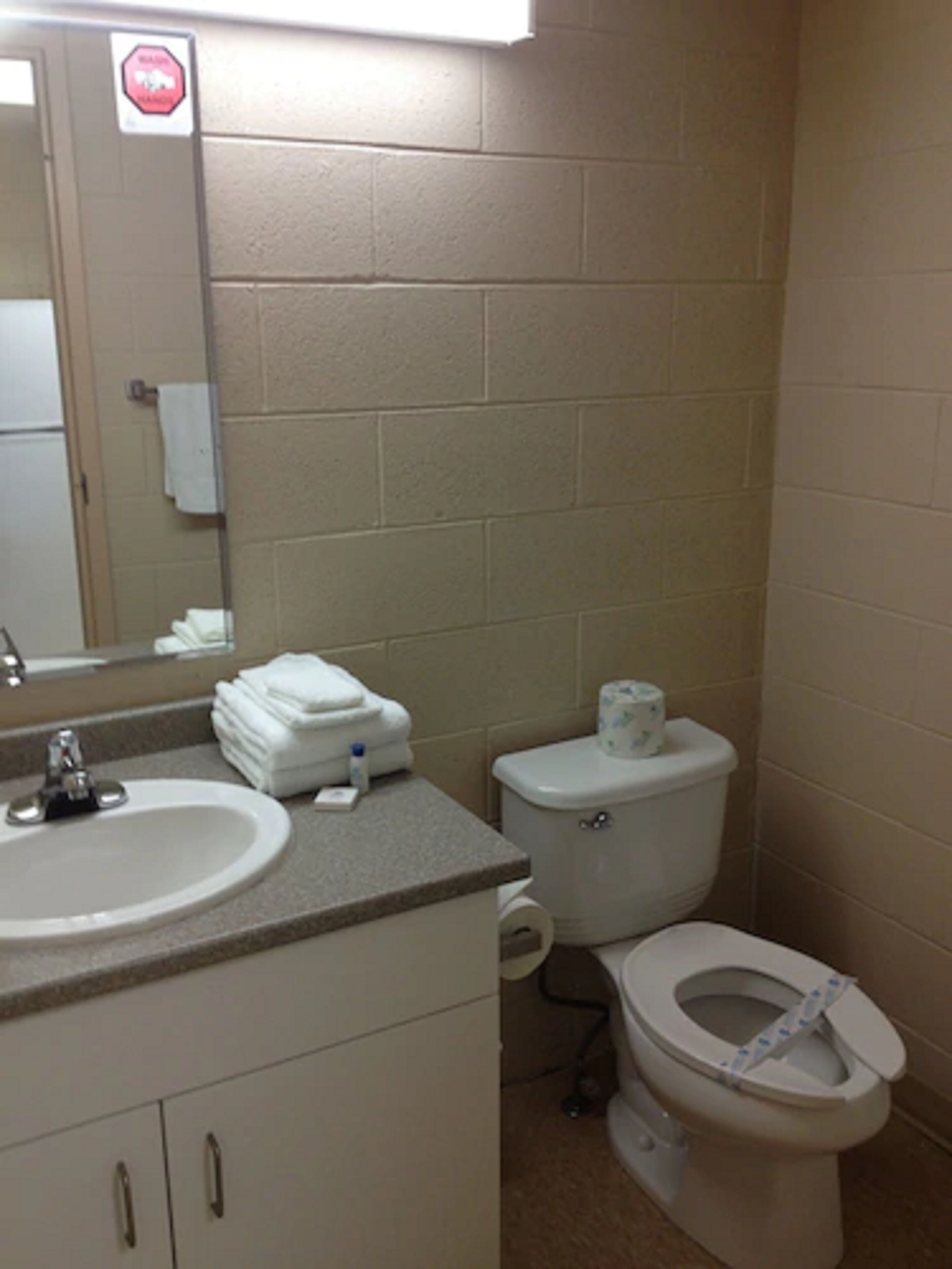 A bathroom with a sink, toilet, and a mirror. There is a stop sign on the wall and a stack of towels on the counter.