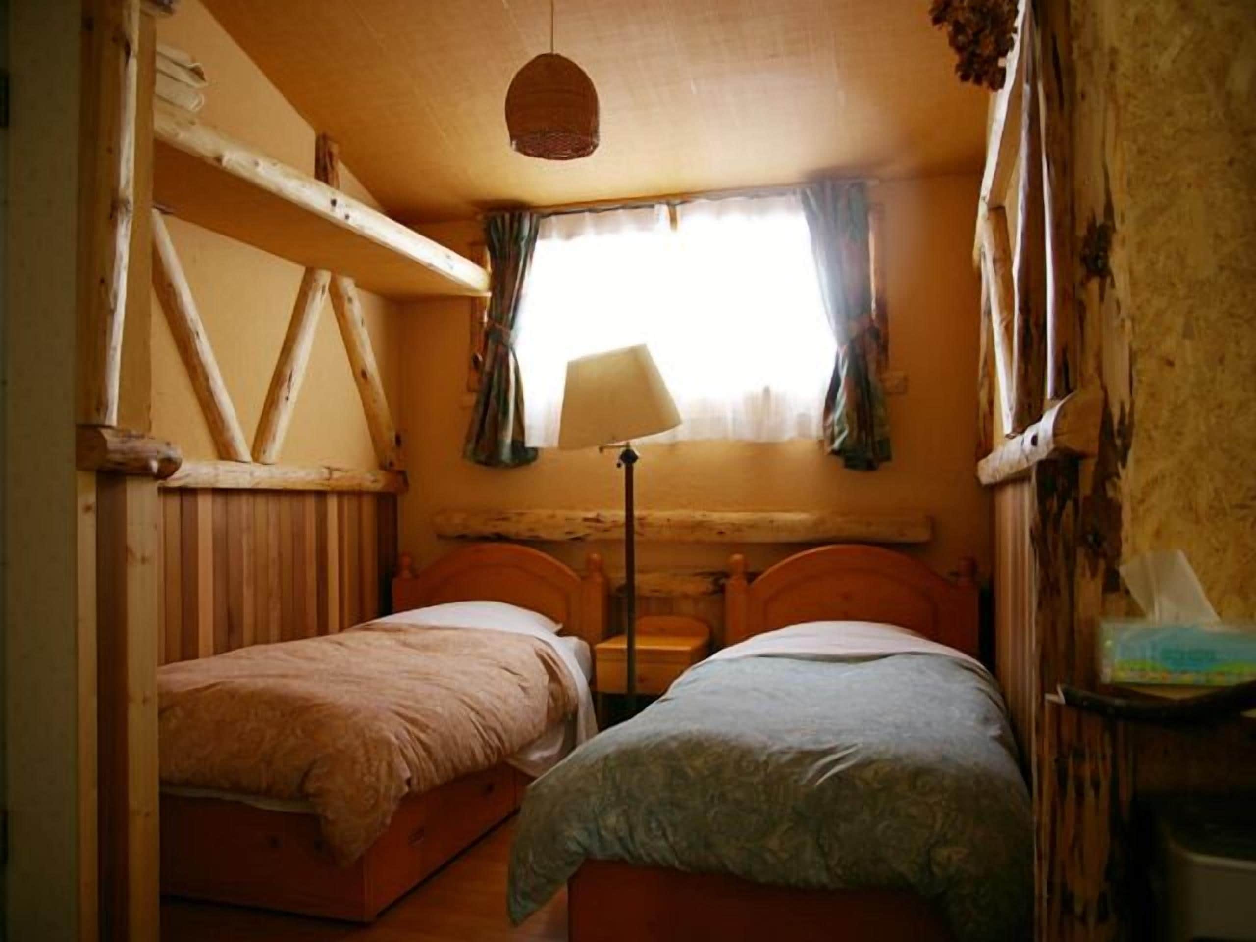 Una habitación pequeña y rústica con dos camas de madera, una ventana con cortinas y una lámpara colgante.