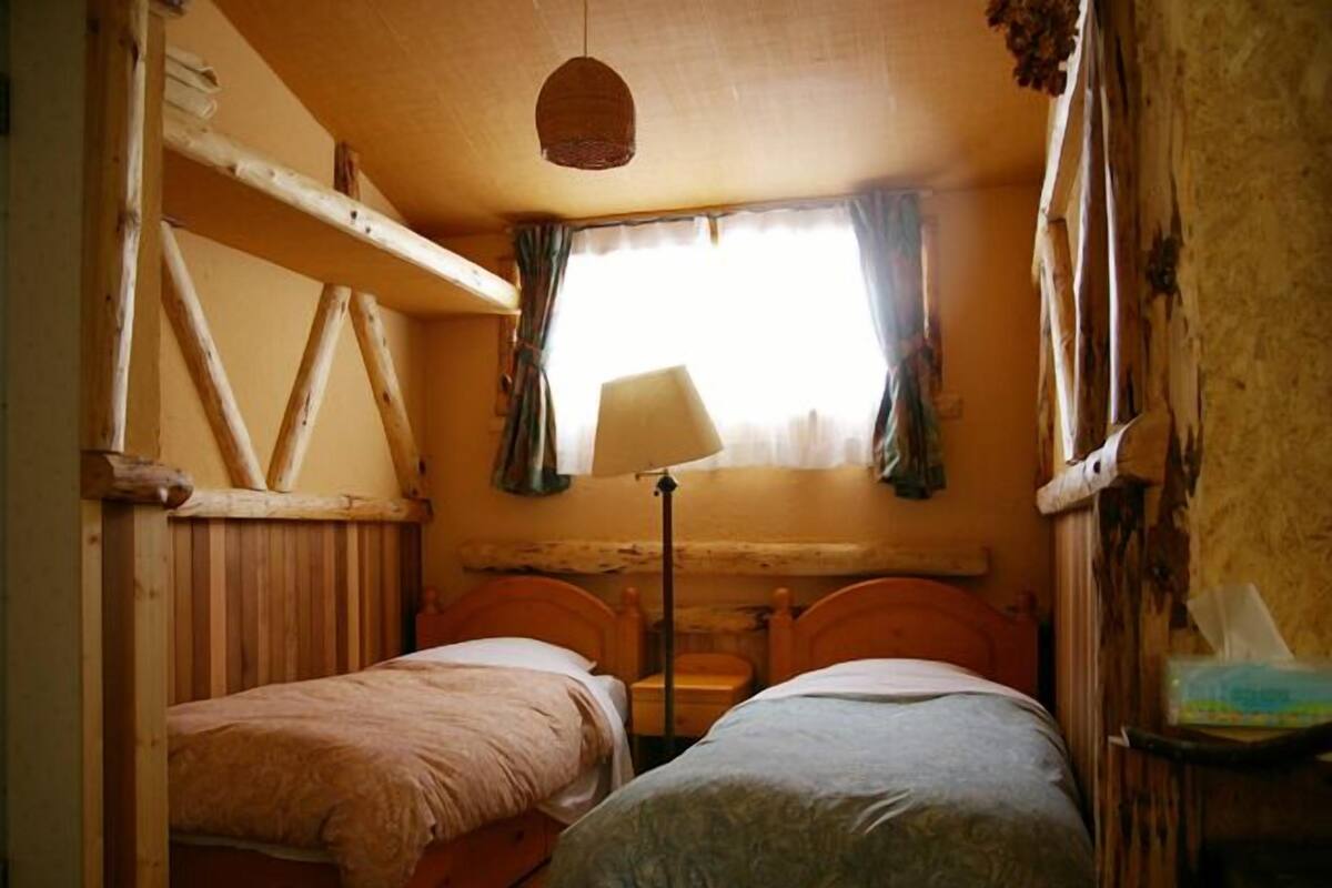 Une petite pièce rustique avec deux lits en bois, une fenêtre simple avec des rideaux et un luminaire suspendu.