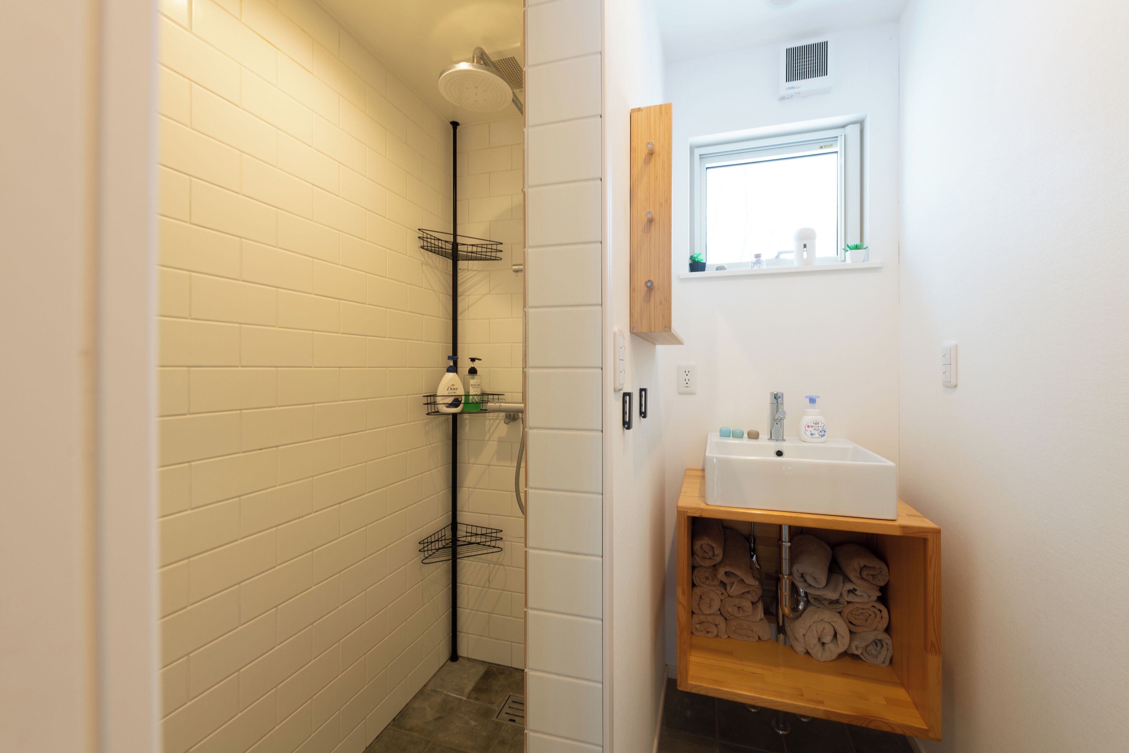 A bathroom with white tiled walls, a wooden shelf with towels, a white sink, and a window above the mirror.