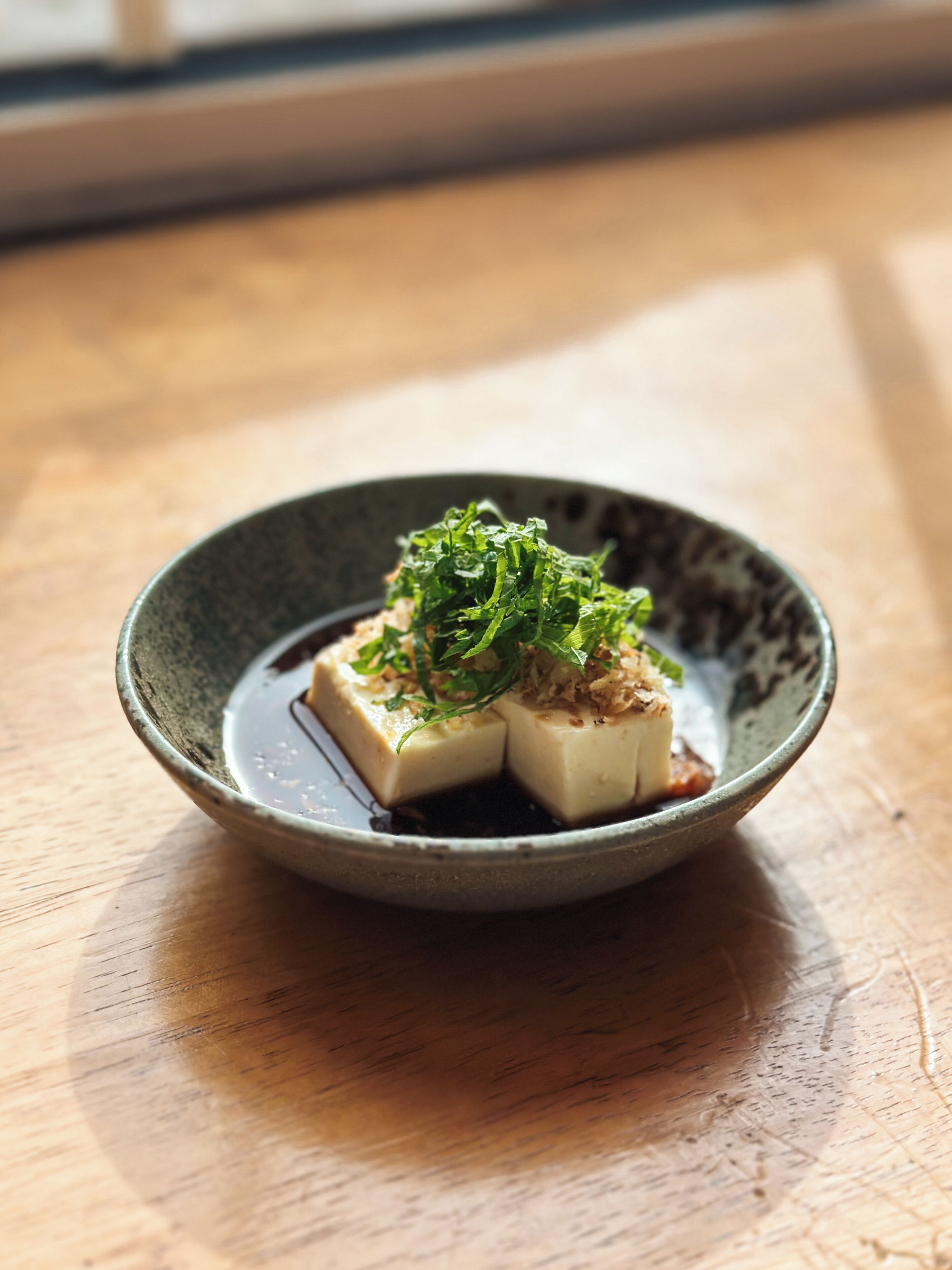 A dish of tofu with green garnish and sauce in a ceramic bowl on a wooden table.