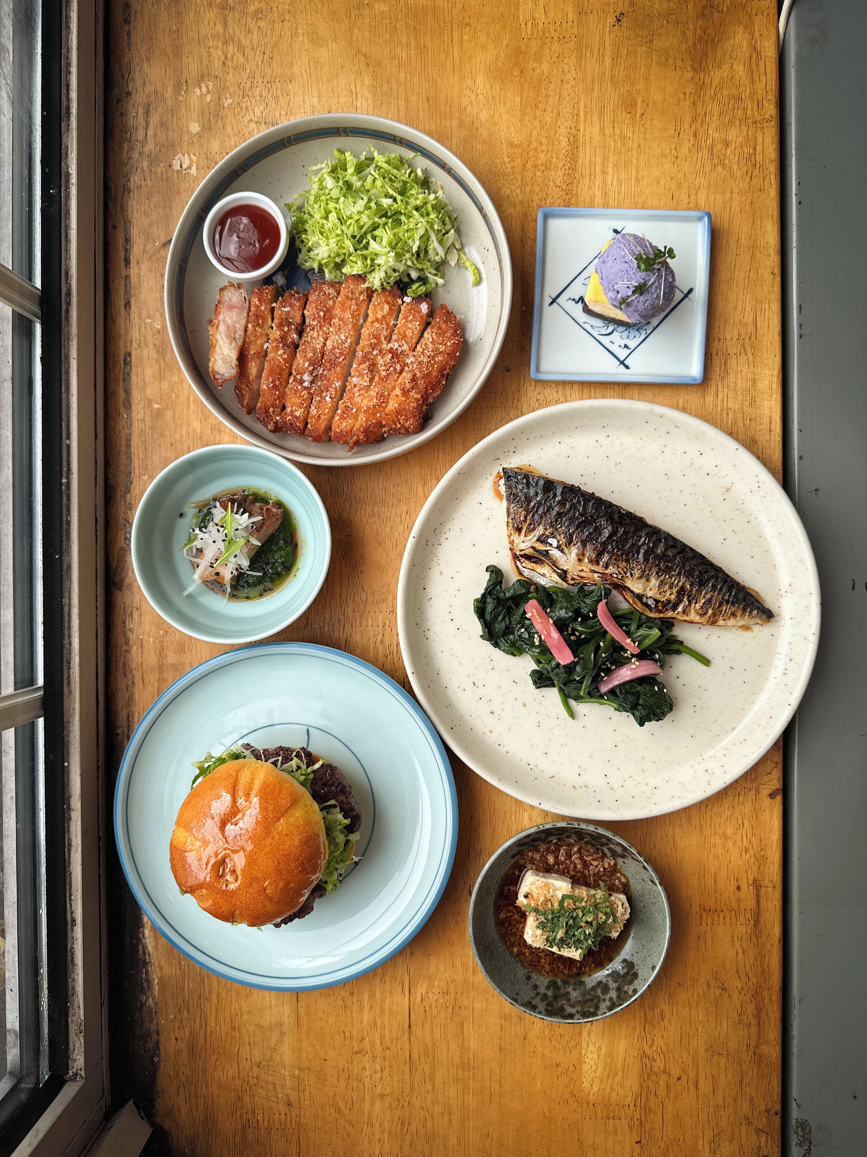 Une table en bois sur laquelle on trouve différents plats, notamment un poisson, un hamburger et une assiette de nourriture frite.