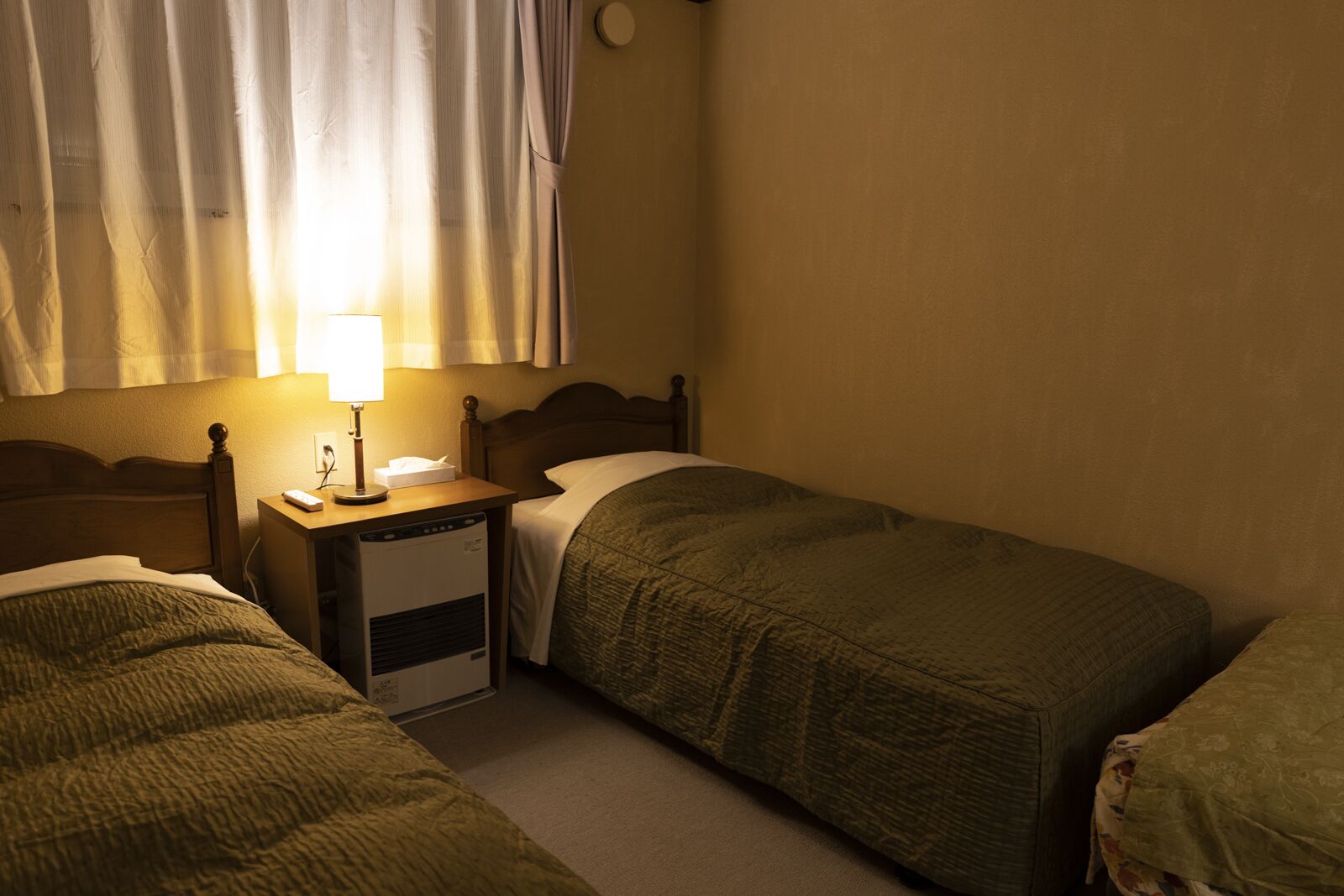 Um quarto pequeno e com pouca luz, com duas camas de solteiro, uma mesa de cabeceira com abajur e um aquecedor.