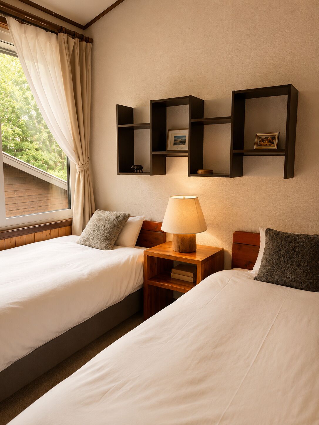 A hotel room with two beds, a nightstand with a lamp, and wall-mounted shelves with pictures.