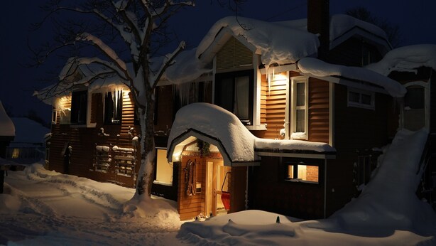 A snow-covered house with a wooden exterior and a lit entrance. - Country Inn Milky House (Niseko)