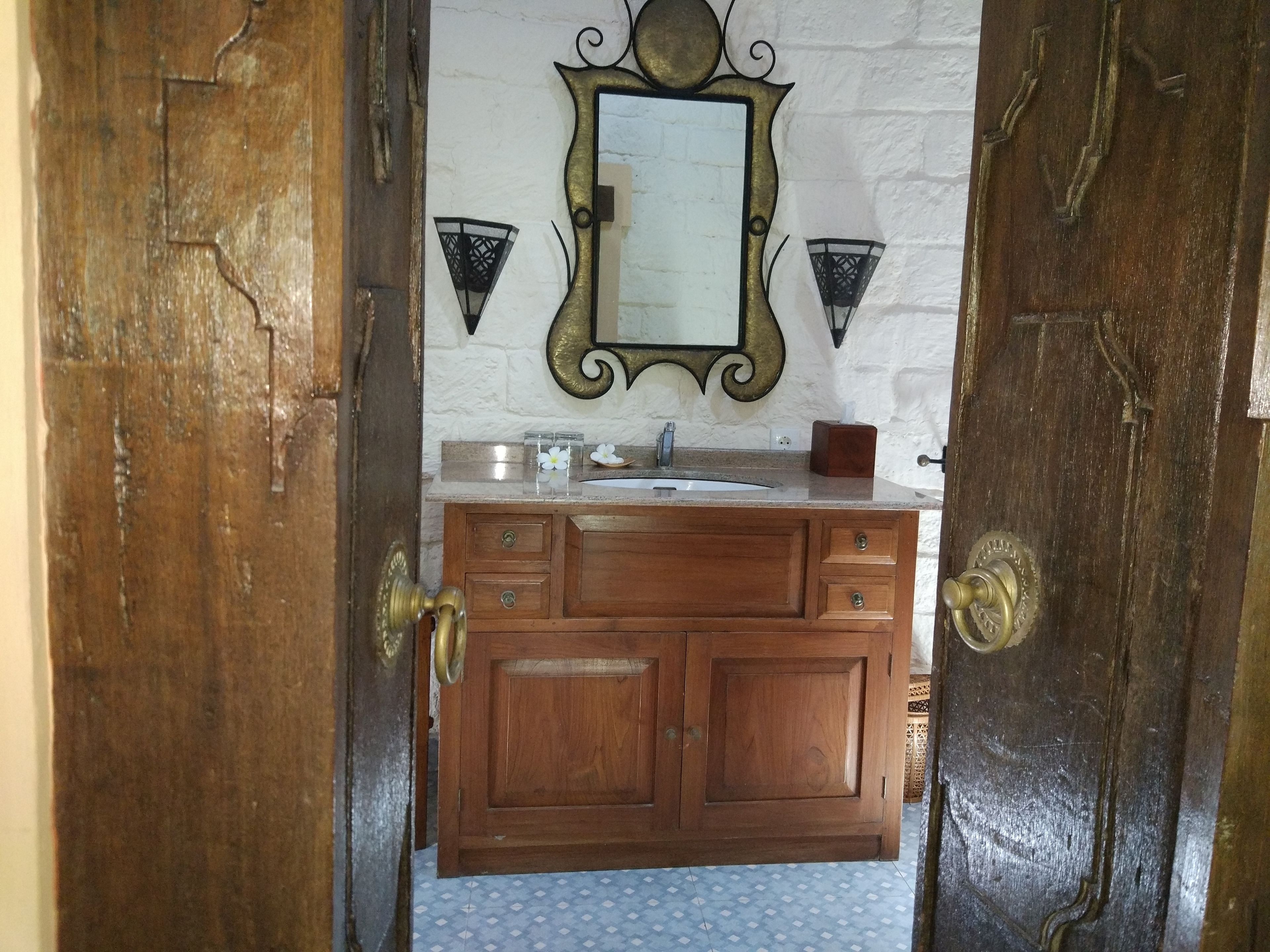 Puerta de madera que da a un baño con suelo blanco de azulejos, un lavabo y un espejo con apliques decorativos en la pared.