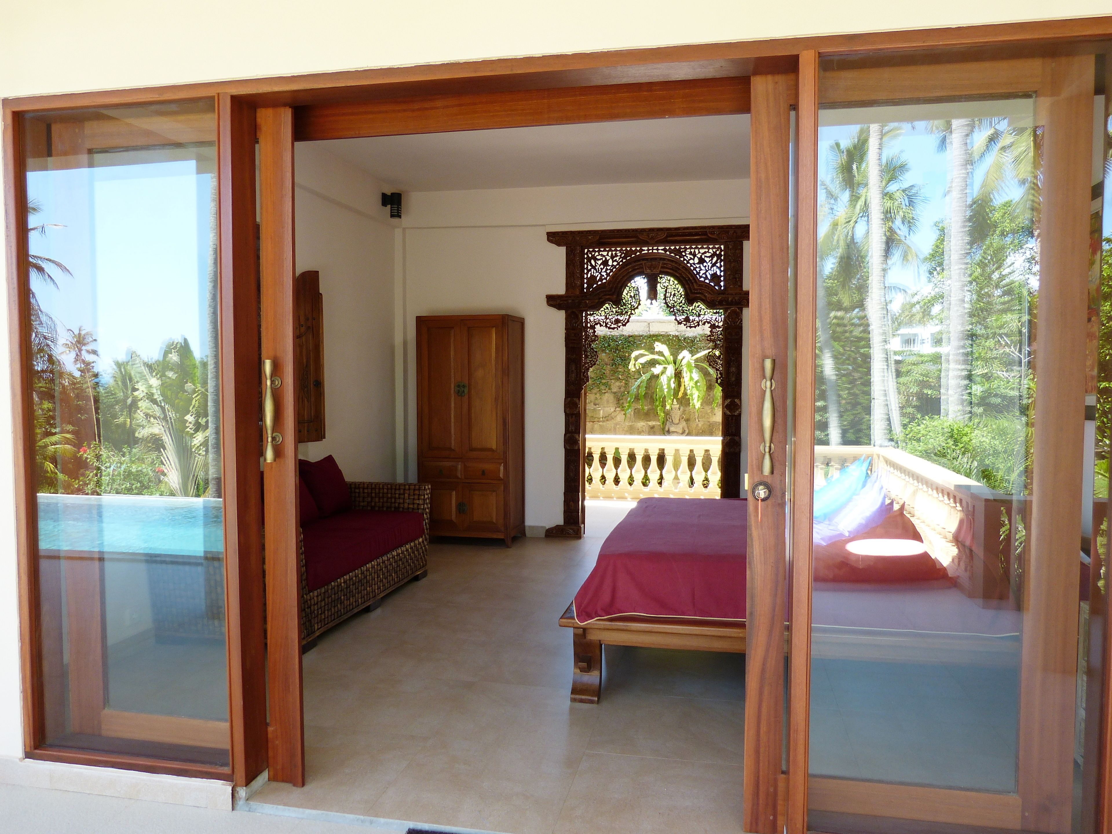 A room with a bed, a wardrobe, and a balcony with a view of palm trees.