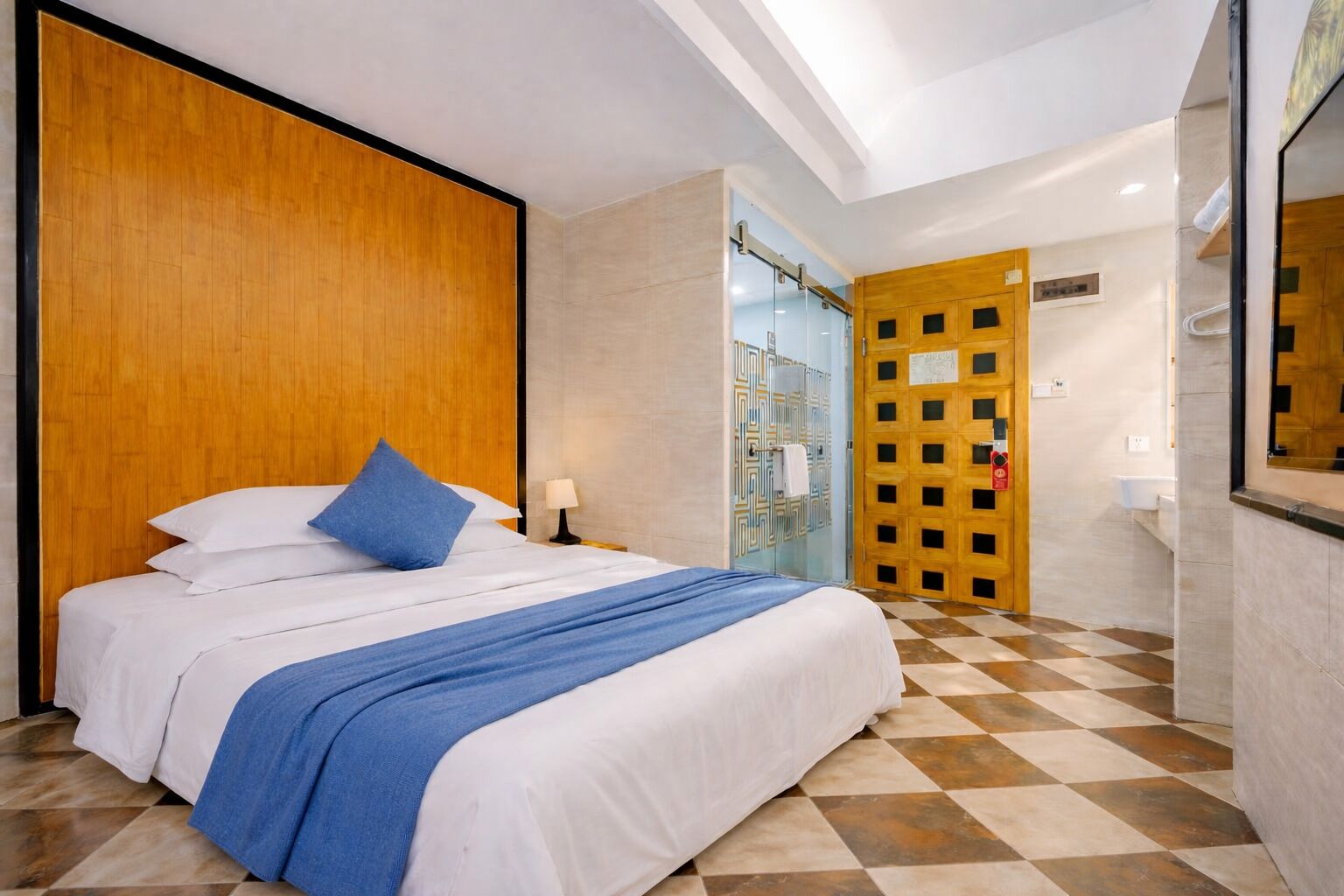 Una camera d'hotel con un letto, una testiera in legno, un pavimento a scacchi, un bagno con porta in vetro e uno specchio.