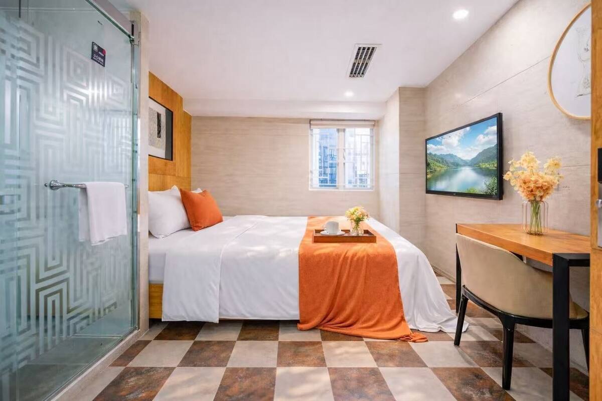 A hotel room with a bed, a desk, a chair, a window, and a shower area.
