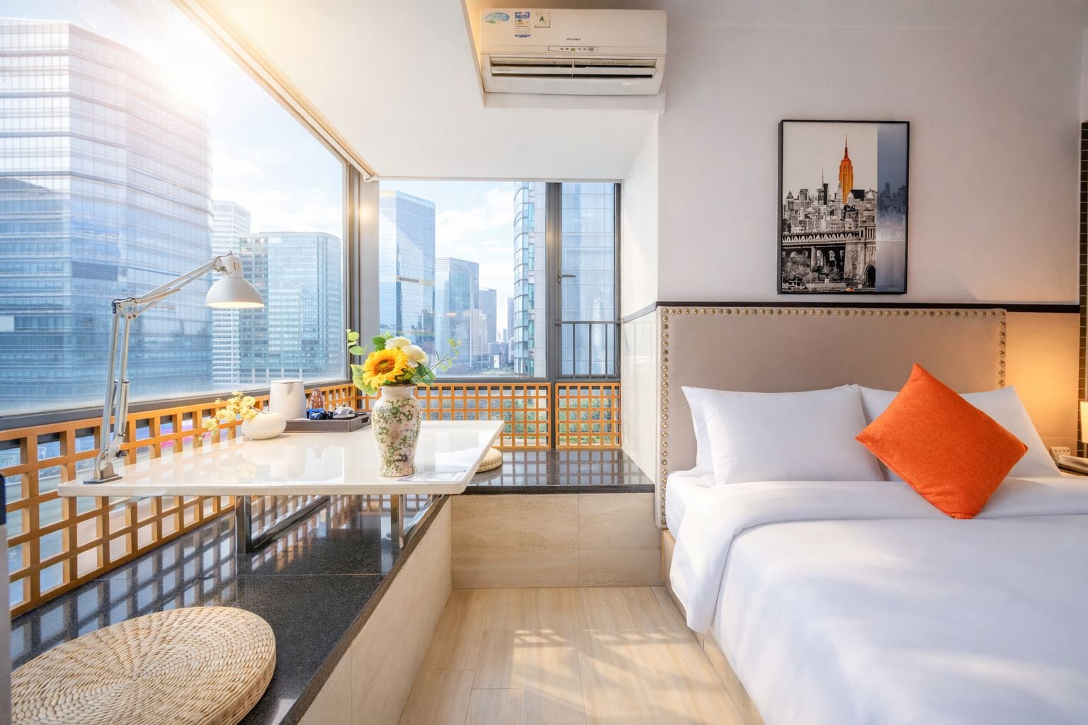 A hotel room with a city view, a desk with a lamp, a vase with flowers, a bed with white bedding and an orange pillow, and a framed picture on the wall.