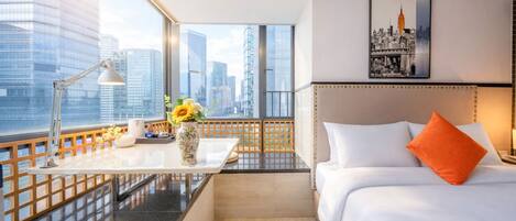 A hotel room with a city view, a desk with a lamp, a vase with flowers, a bed with white bedding and an orange pillow, and a framed picture on the wall.