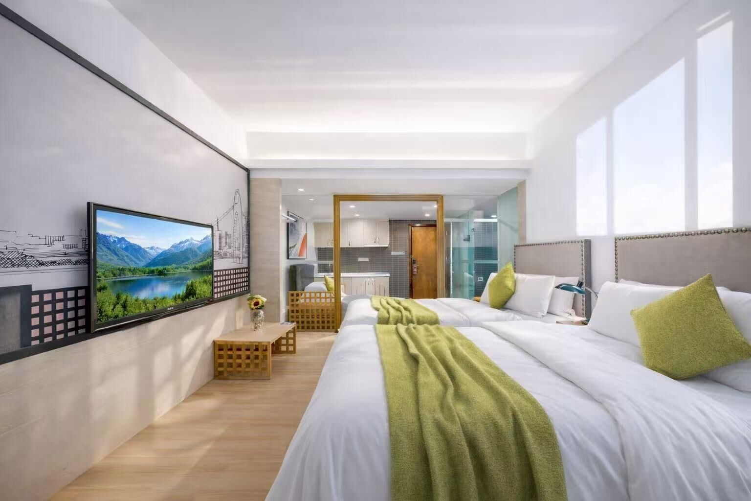 A modern hotel room with a large bed, a TV, a view of a mountain landscape, and a kitchenette in the background.