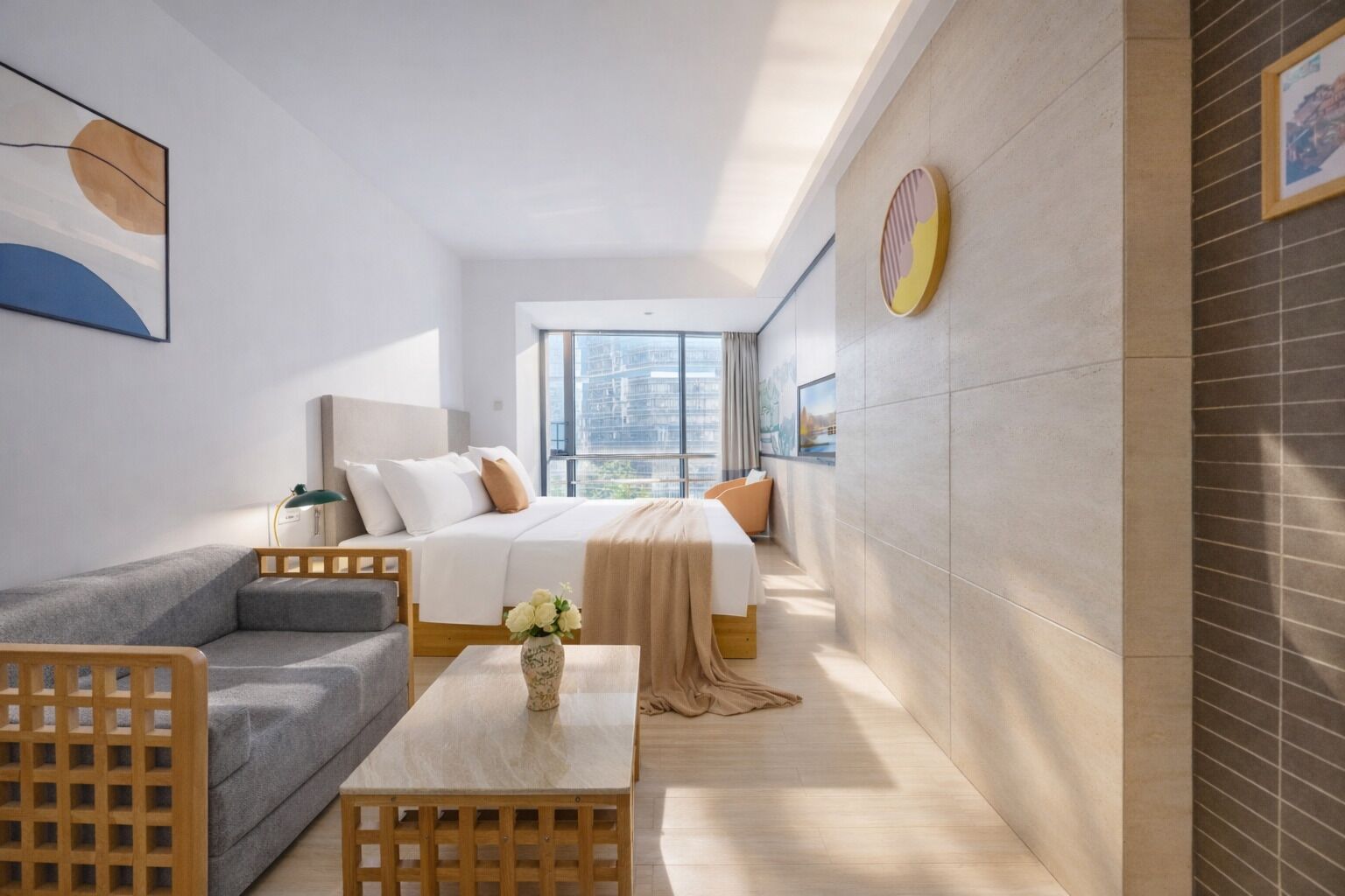 A modern hotel room with a bed, a sofa, a coffee table, and a large window offering a city view.