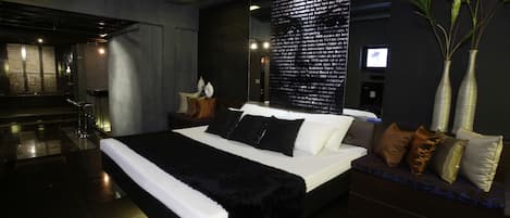 A modern hotel bedroom with a large bed, bedside tables, a nightstand, a TV, and a wall-mounted screen.
