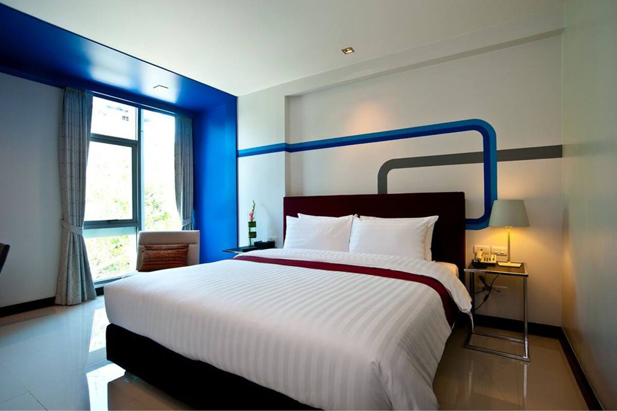 A modern hotel room with a large bed, a nightstand, a lamp, and a view of greenery through the window.