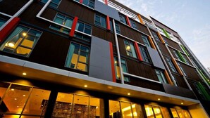 Un hôtel à plusieurs étages doté d’une architecture moderne, caractérisé par un mélange de panneaux de bois foncés et clairs, de grandes fenêtres et une façade où les couleurs rouge et verte sont mises en valeur.
