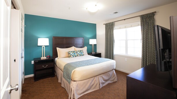 A bedroom with a bed, nightstands, a television, and a window with curtains. - The Colonies at Williamsburg (Williamsburg)