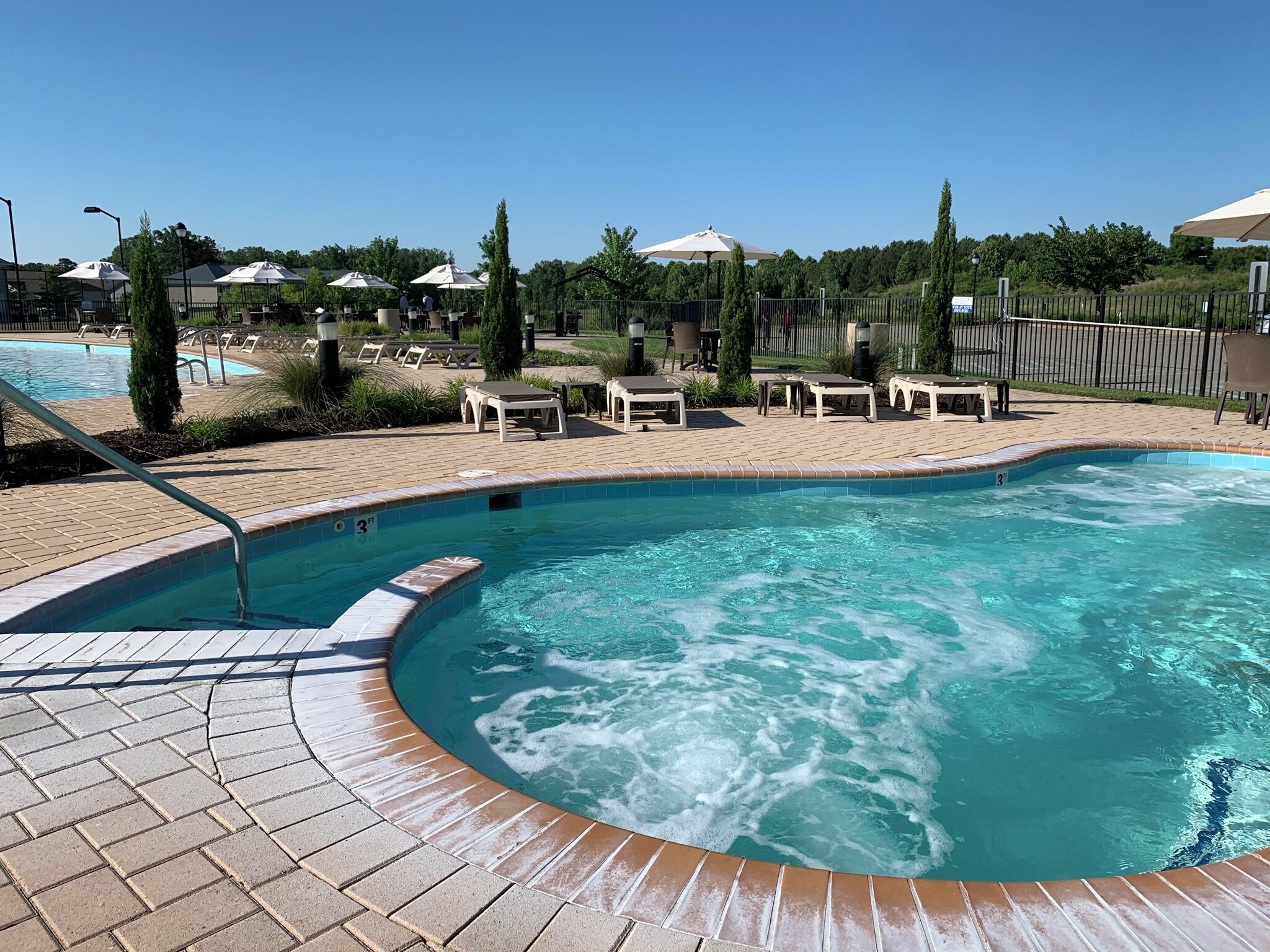 A swimming pool with a hot tub, surrounded by lounge chairs and umbrellas, with a brick patio and greenery in the background.