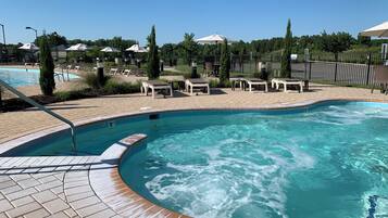 Una piscina con hidromasaje, rodeada de sillones de descanso y sombrillas, con un patio de ladrillo y follaje al fondo.