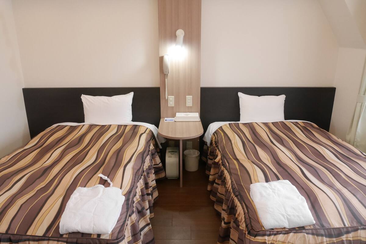 Ett hotellrum med två sängar, båda med randiga sängkläder och vita kuddar. Mellan sängarna står ett litet bord med en lampa, och bredvid finns en papperskorg.