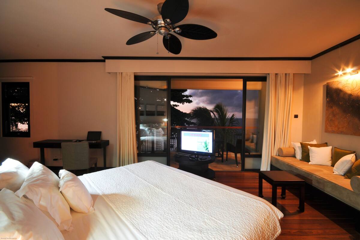 A hotel room with a large bed, a desk with a computer, a TV, and a balcony with a view of the evening sky.