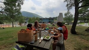 Un grupo de personas cenando en una mesa al aire libre con vistas a una montaña y edificios modernos al fondo.