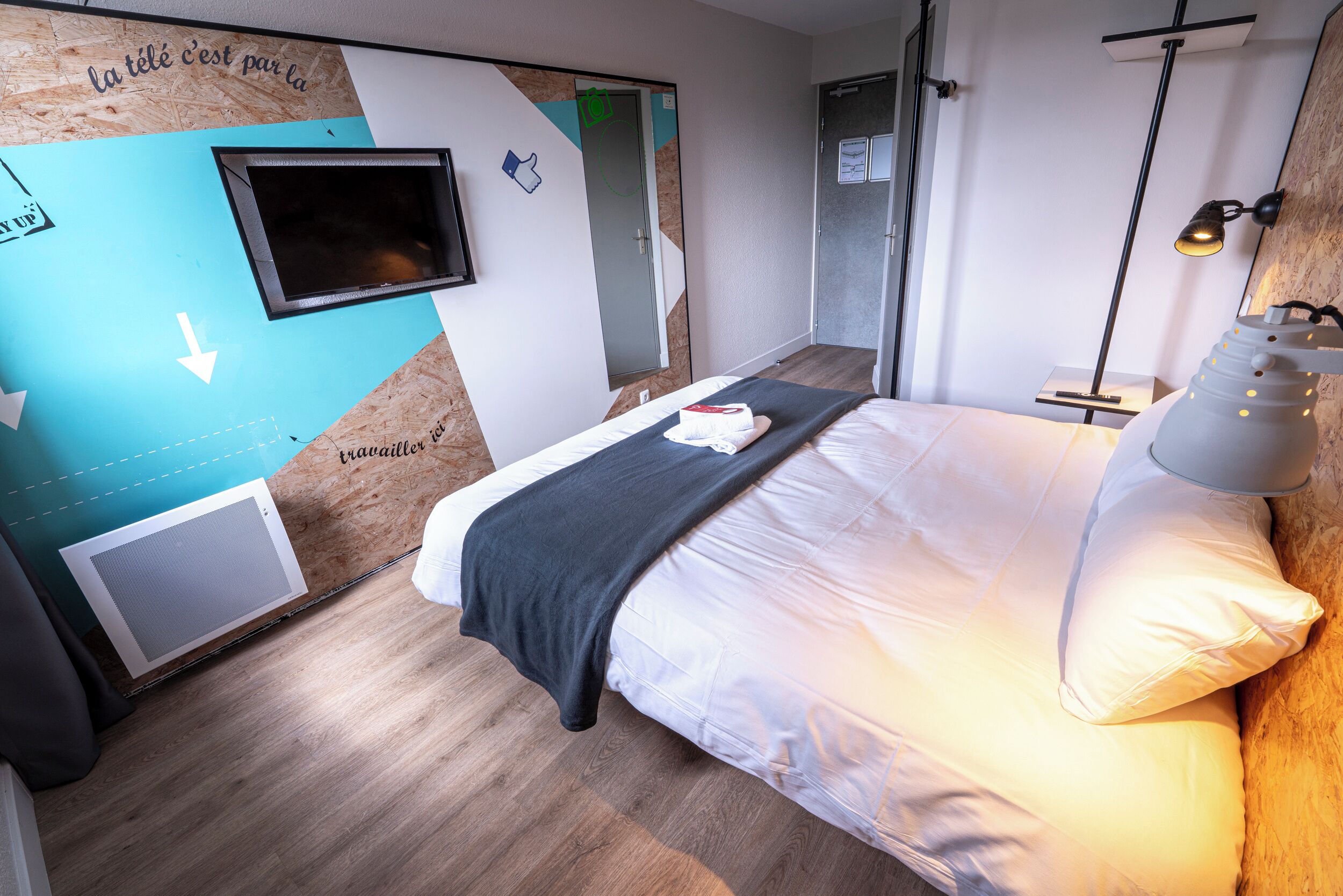 A modern hotel room with a bed, a TV, a desk, and a lamp.