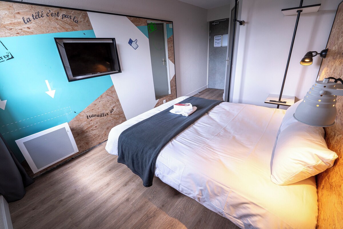 A modern hotel room with a bed, a TV, a desk, and a lamp.