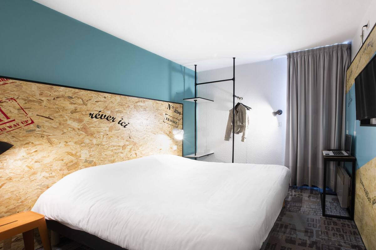 A modern hotel room with a large bed, a wooden headboard with text, a small desk with a laptop, a hanging coat rack, and a TV mounted on the wall.